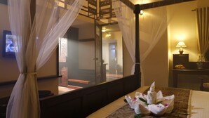 Ein Zimmer mit einem Himmelbett, einem Fernseher, einem Schreibtisch mit Lampe und Blick ins Freie.