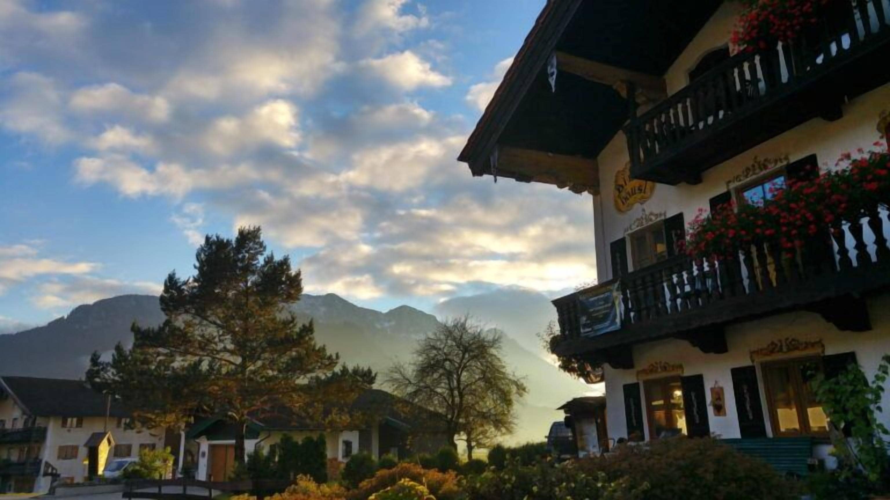 A traditional chalet-style building with a balcony and flower boxes, surrounded by trees and mountains in the background.