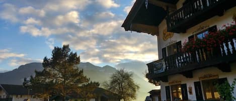 A traditional chalet-style building with a balcony and flower boxes, surrounded by trees and mountains in the background.