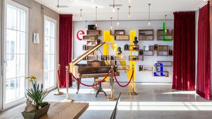 Un piano à queue se trouve dans une pièce spacieuse, dotée de grandes fenêtres, de rideaux en velours rouge et de plusieurs étagères où sont disposés des livres et des objets décoratifs.
