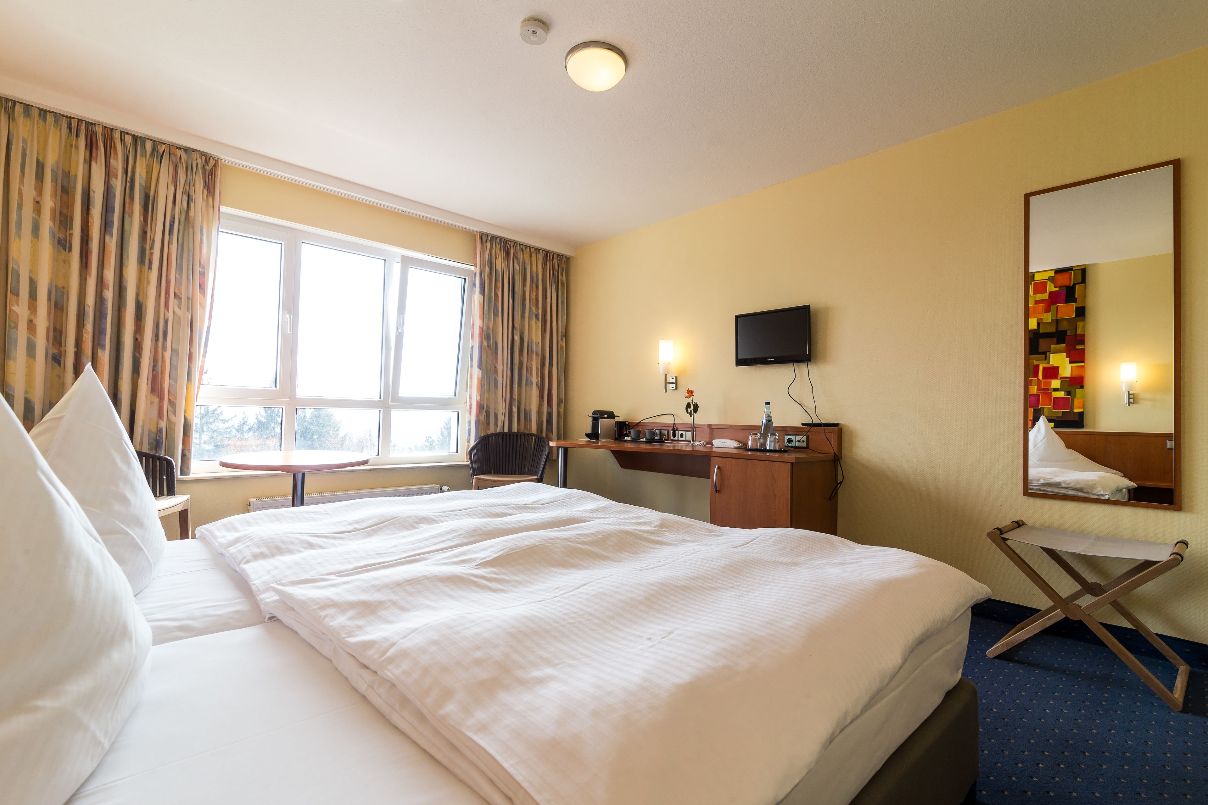 A hotel room with a large bed, a desk, a chair, a small table, a television, and a window with curtains.