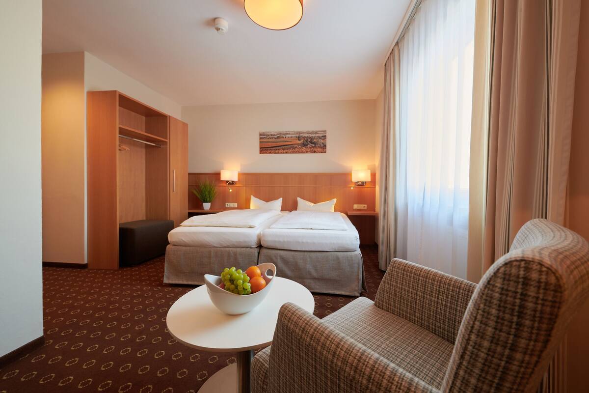 Une chambre d’hôtel avec un lit double, deux tables de chevet avec des lampes, une petite table ronde avec un panier de fruits, un canapé et une armoire.