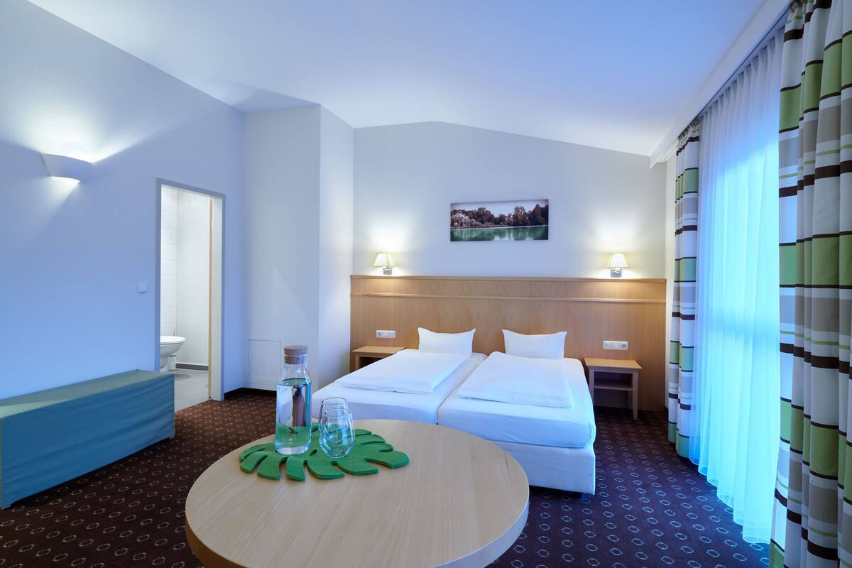 Une chambre d’hôtel avec deux lits, une table ronde en bois surmontée d’une nappe verte, un tableau accroché au mur et une fenêtre avec des rideaux.