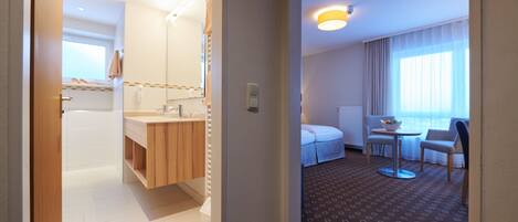 A hotel room with a bathroom, a bed, a small table with a bowl of fruit, and a window with curtains.