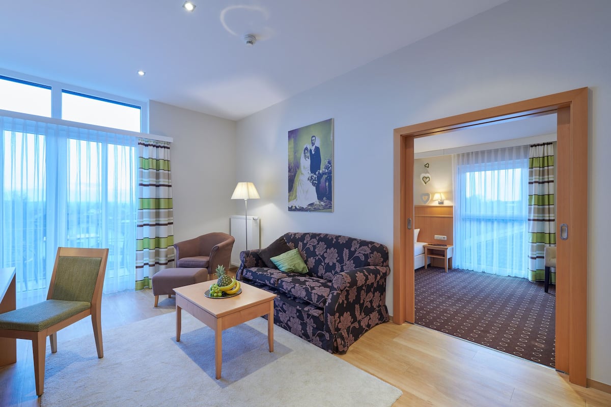Un salon moderne avec un canapé, un fauteuil et une table basse. Un tableau est accroché au mur et, par une porte ouverte, on aperçoit une autre pièce.