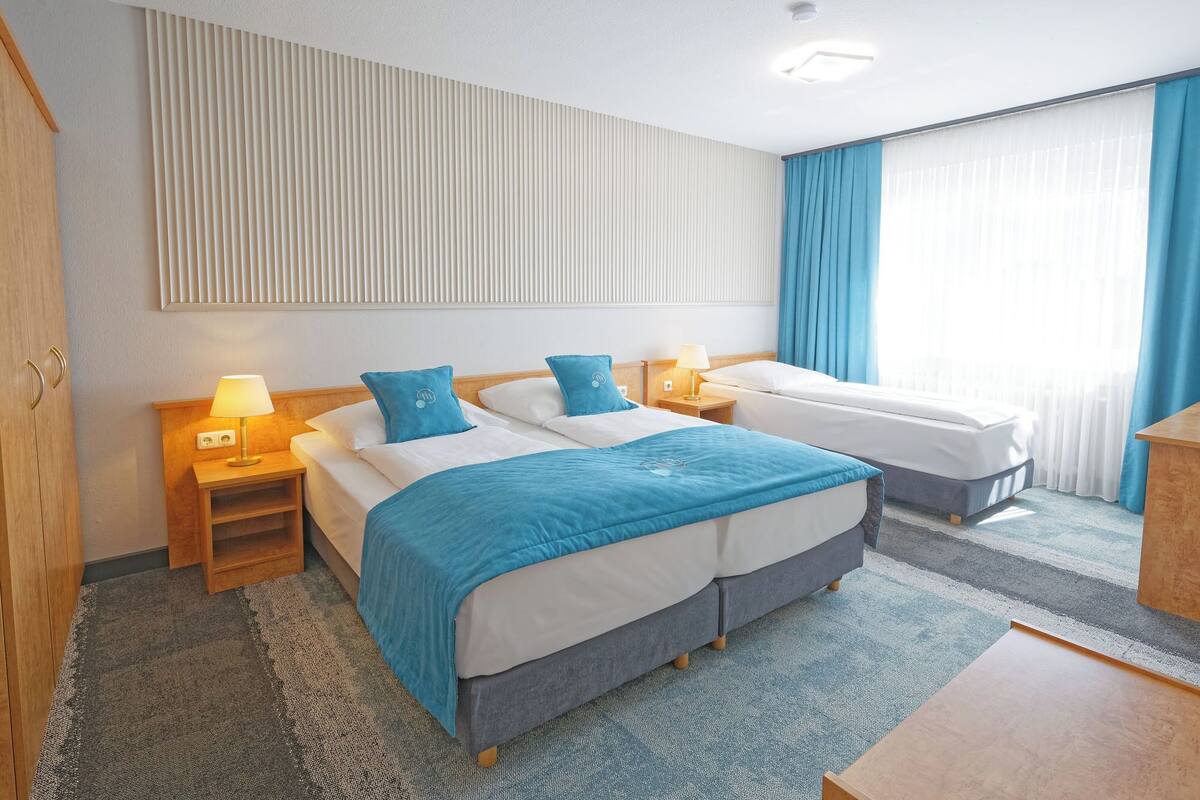 A hotel room with two beds, a wooden wardrobe, a nightstand with a lamp, and a window with curtains.