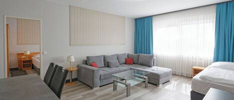 A modern hotel room with a grey sofa, a glass coffee table, a wooden side table, and a bed with white bedding.