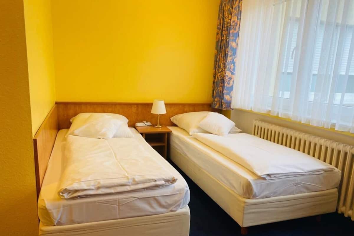 Two single beds with white linens, a bedside table with a lamp, a radiator, and a window with sheer curtains.