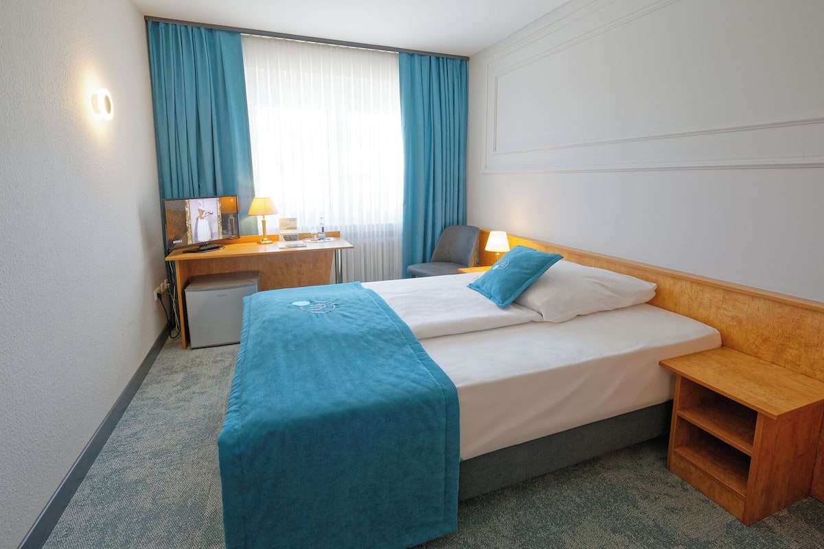 A hotel room with a bed, a desk, a chair, a small refrigerator, and a nightstand.