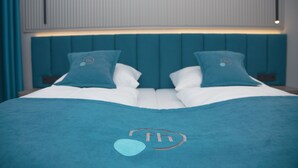 A neatly made bed with a teal headboard and pillows, a teal blanket with a logo, and a window with blinds in the background. - Friends Hotel Kerpen (Kerpen)