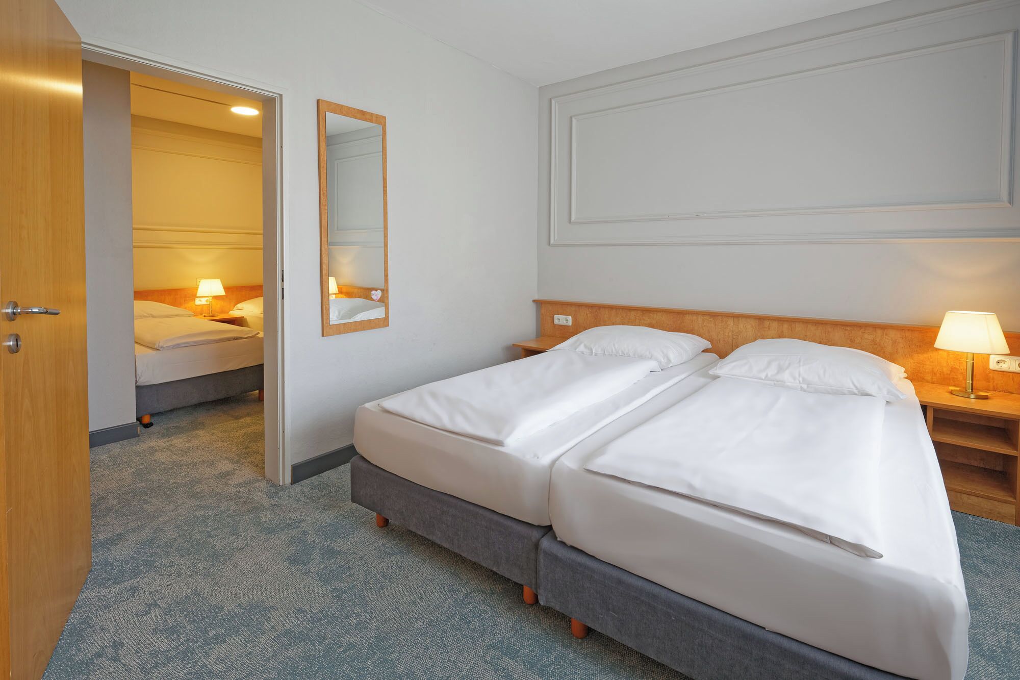 Une chambre d’hôtel avec un lit double, une table de chevet avec une lampe, un miroir et une porte donnant sur une autre pièce.