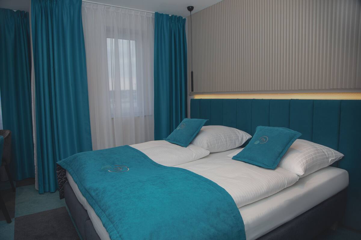 A neatly made bed with teal bedding and pillows, a headboard with vertical paneling, and a window with sheer white curtains.