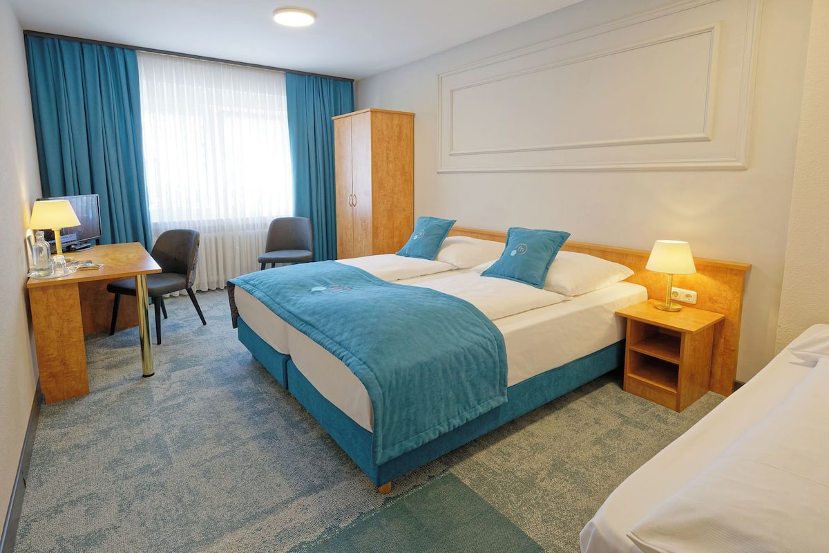 A hotel room with a bed, a desk with a lamp, a chair, a wardrobe, and a television.