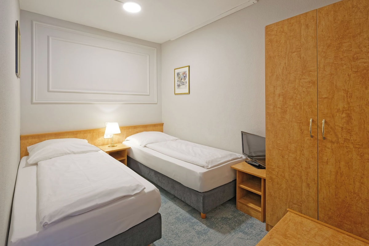 A hotel room with two beds, a wooden wardrobe, a desk with a lamp, and a framed picture on the wall.