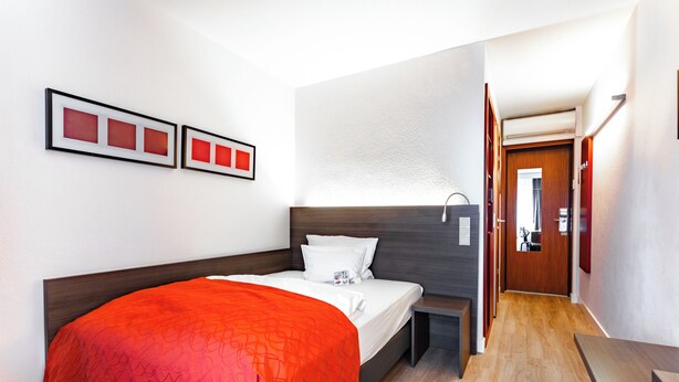 Una habitació d'hotel amb un llit, una taula de nit, una làmpada penjada a la paret i una porta que dona a una altra estança.