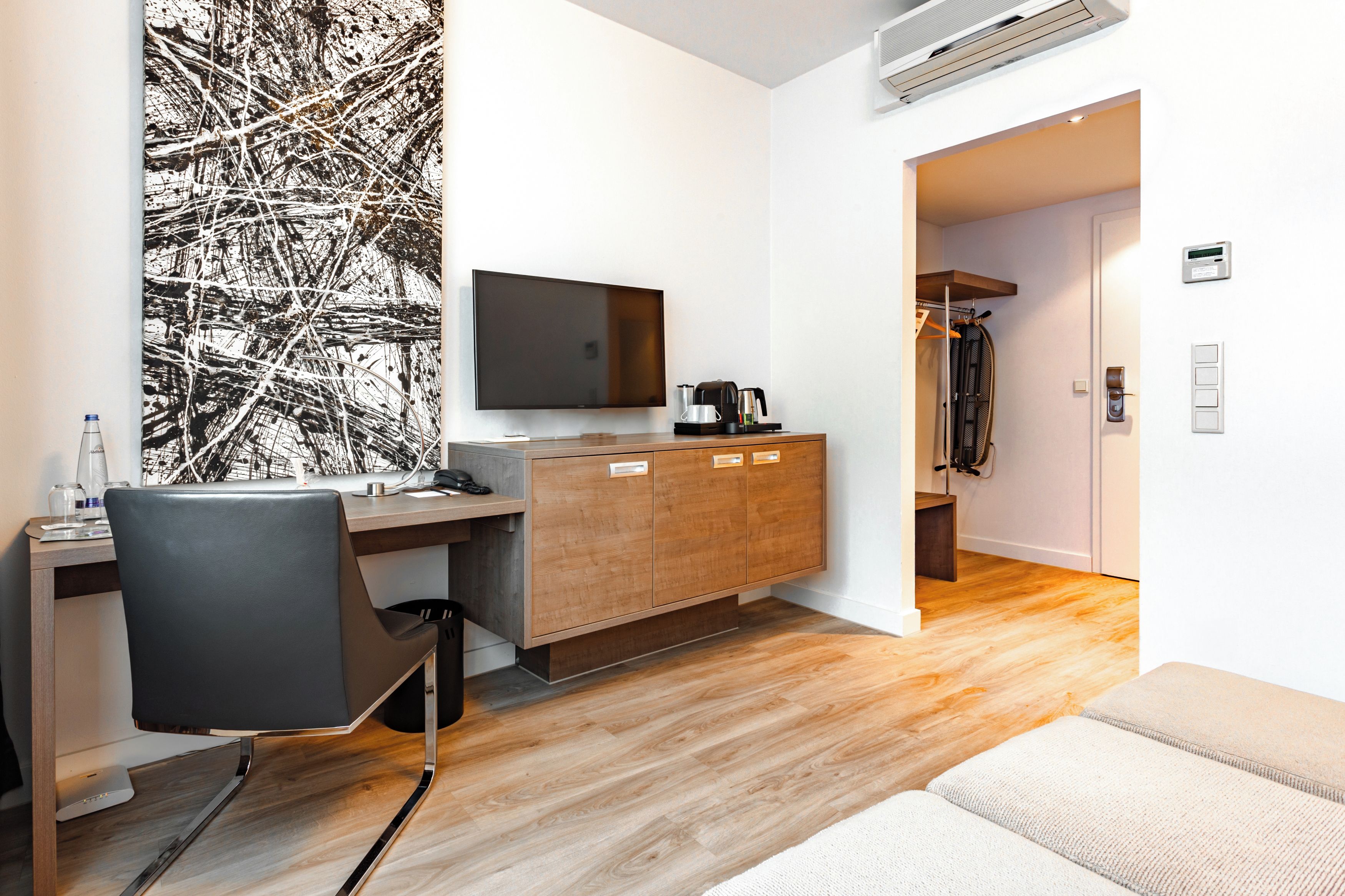 A modern hotel room with a wooden desk, a black office chair, a flat-screen TV, a coffee maker, and a wardrobe.