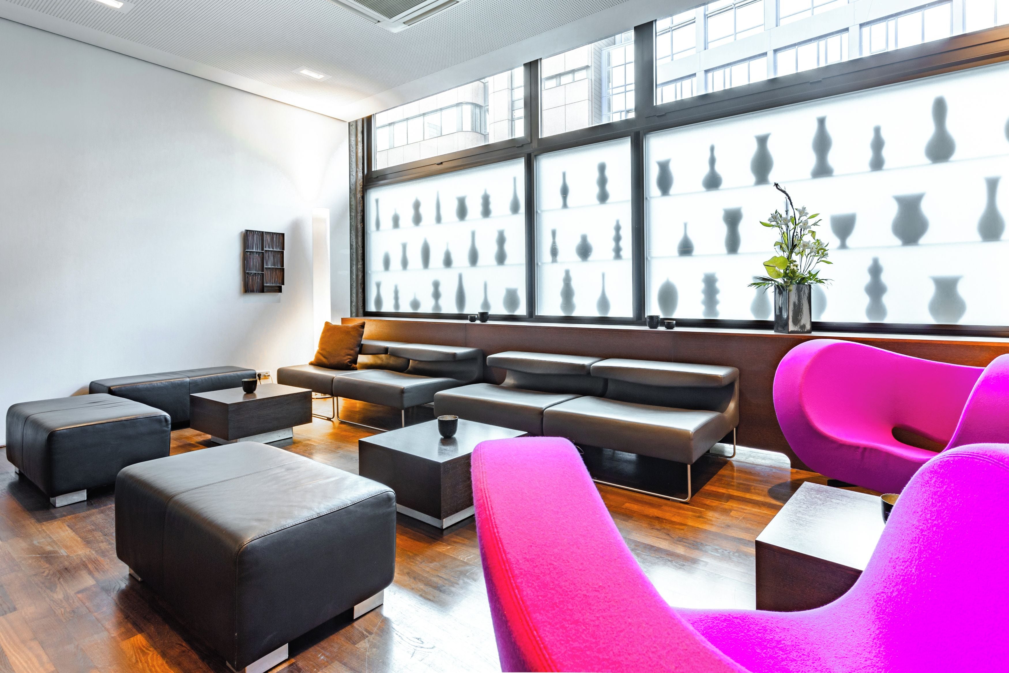 Un salón moderno con asientos negros y rosados, un ventanal con siluetas de jarrones y una pequeña planta sobre una mesa.