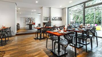 Un comedor moderno con suelo de madera, sillas negras y mesas. Amplias ventanas ofrecen vistas a árboles y una estructura amarilla en el exterior.