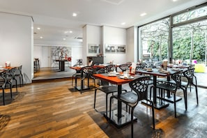 Un comedor moderno con suelo de madera, sillas negras y mesas. Amplias ventanas ofrecen vistas a árboles y una estructura amarilla en el exterior.