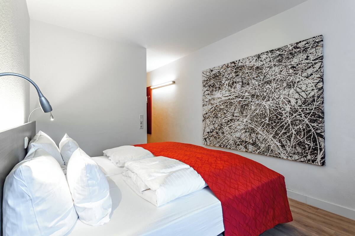 A modern hotel room with a neatly made bed, a large abstract painting on the wall, and a bedside lamp.