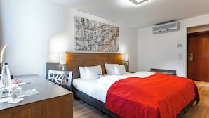 Habitació d'hotel moderna amb un panell de capçalera de fusta, un quadre abstracte gran, un edredó vermell i un escriptori amb una làmpada i una ampolla d'aigua.