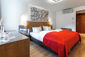 A modern hotel room with a wooden headboard, a large abstract painting, a red bedspread, and a desk with a lamp and a water bottle. - Art Business Hotel Nürnberg (Nuremberg)