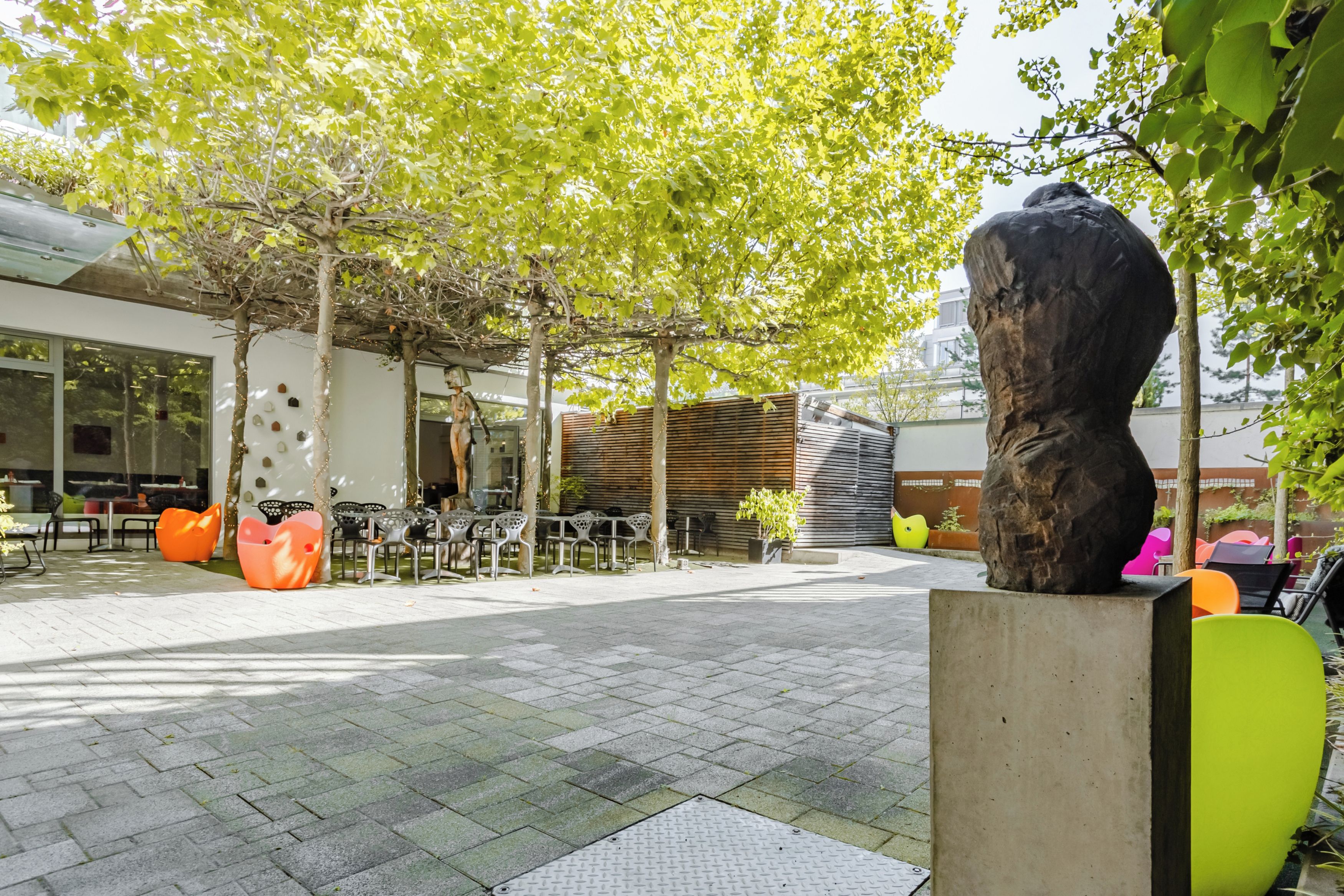 Un espacio al aire libre con sillas y mesas modernas, rodeado de árboles y una gran escultura.