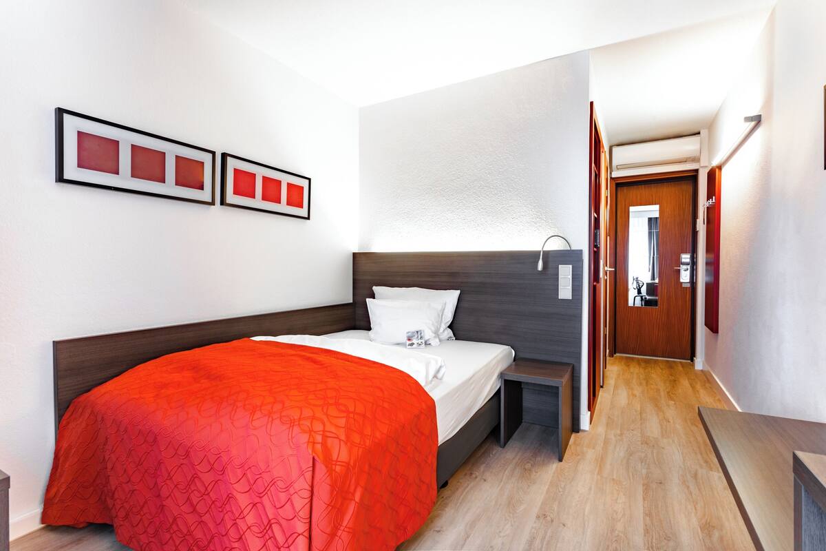 A hotel room with a bed, a bedside table, a wall-mounted light, and a door leading to another room.