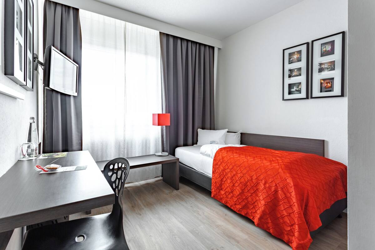 A hotel room with a bed, a desk, a chair, a window with curtains, and framed pictures on the wall.