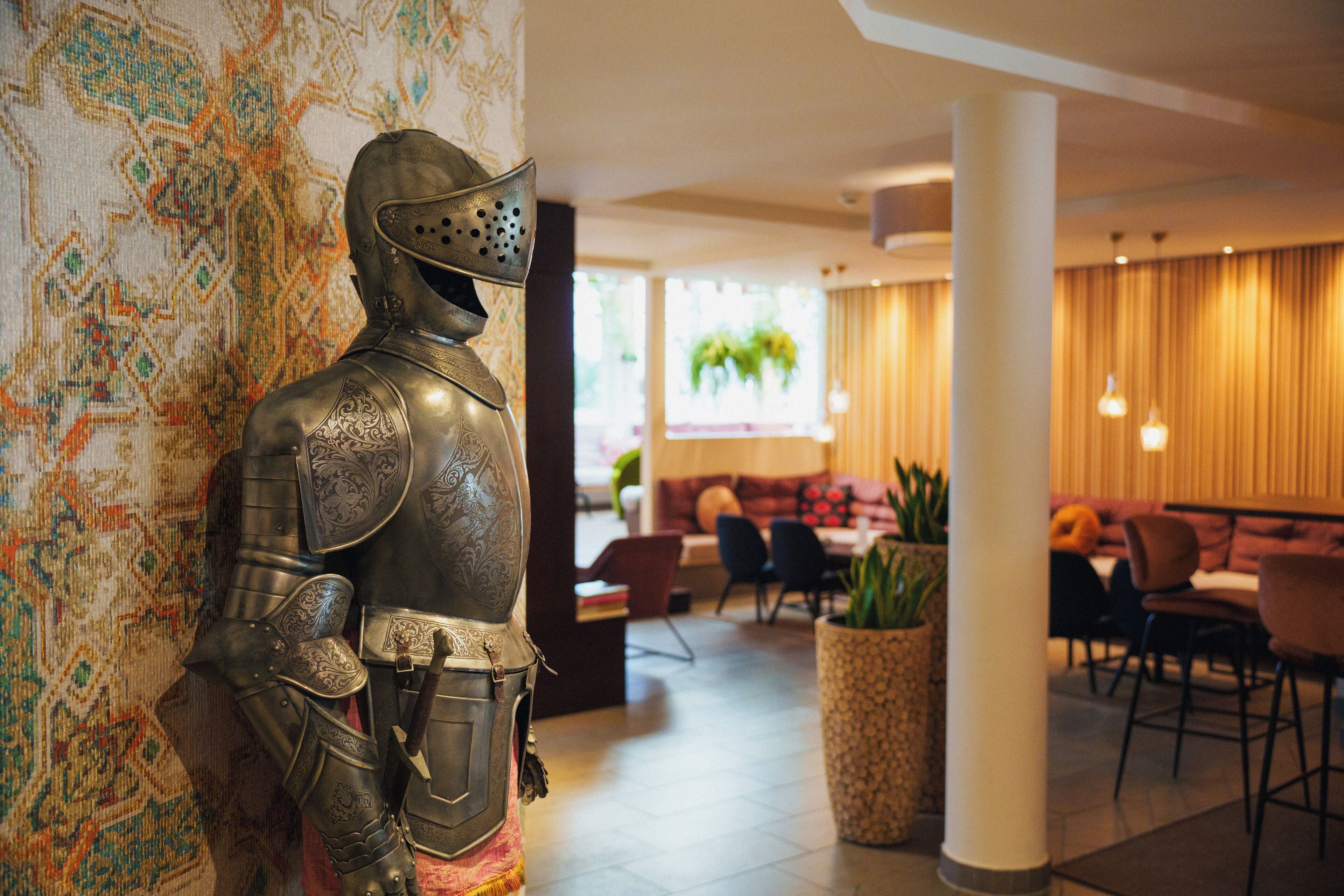 A statue of a knight in armor in a lobby with a patterned carpet, a large pillar, and a seating area with tables and chairs.