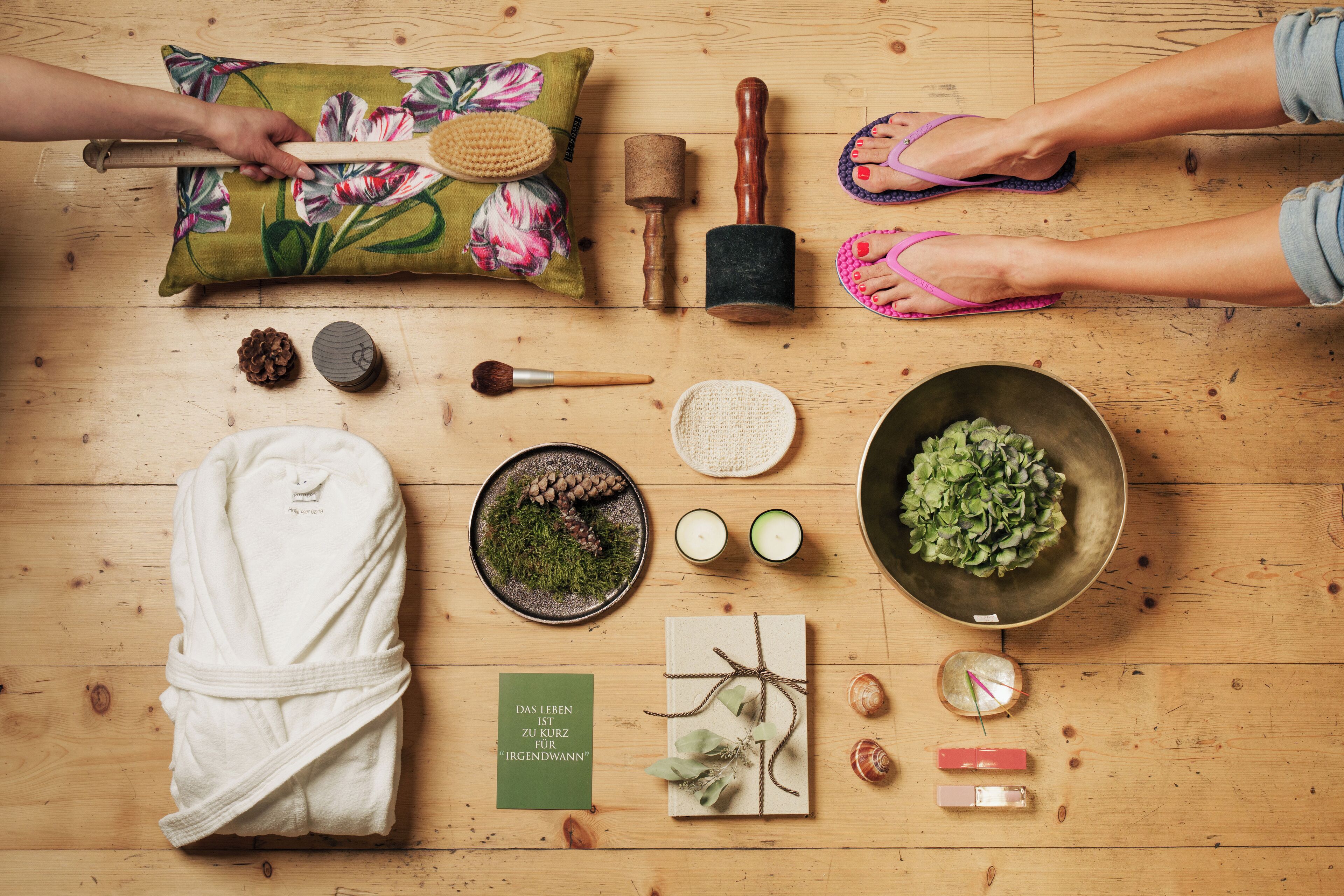 Un plancher en bois, une serviette, une brosse en bois, un maillet en bois, un bol d’herbes aromatiques, un bol de pommes de pin, une bougie, une horloge, une carte et une paire de tongs.