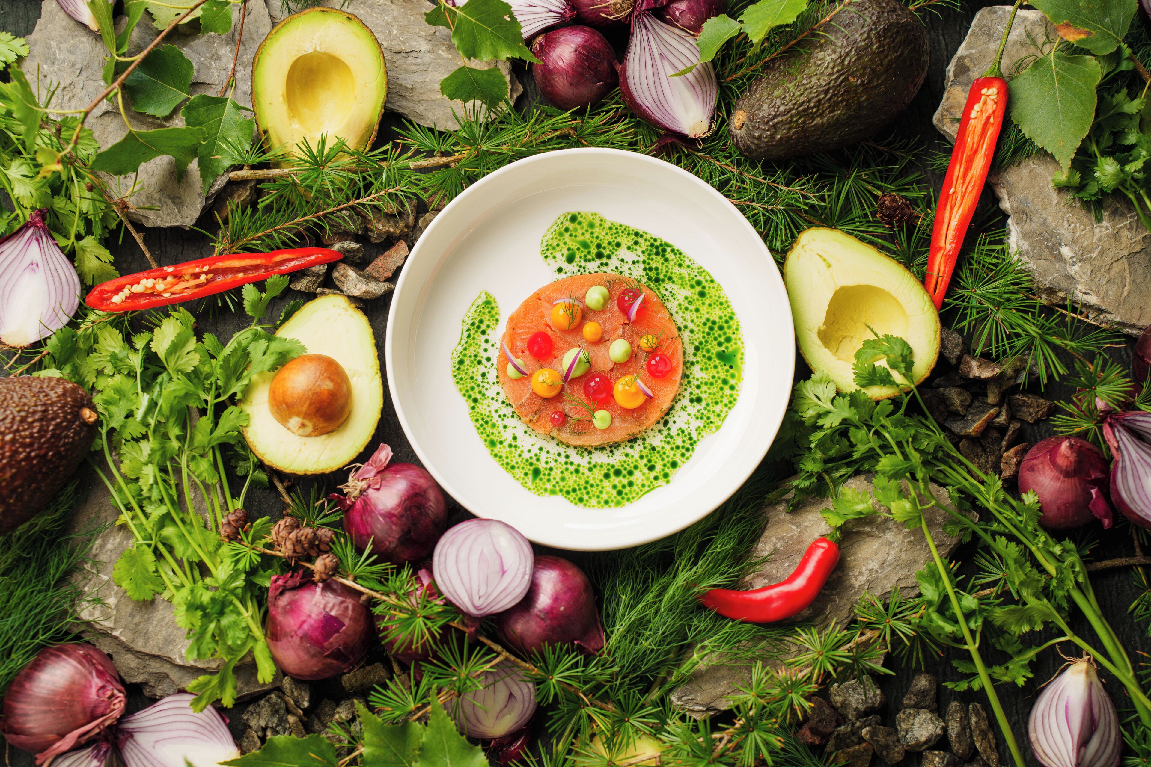 A white bowl with a green sauce and a salmon slice, surrounded by various vegetables and herbs.