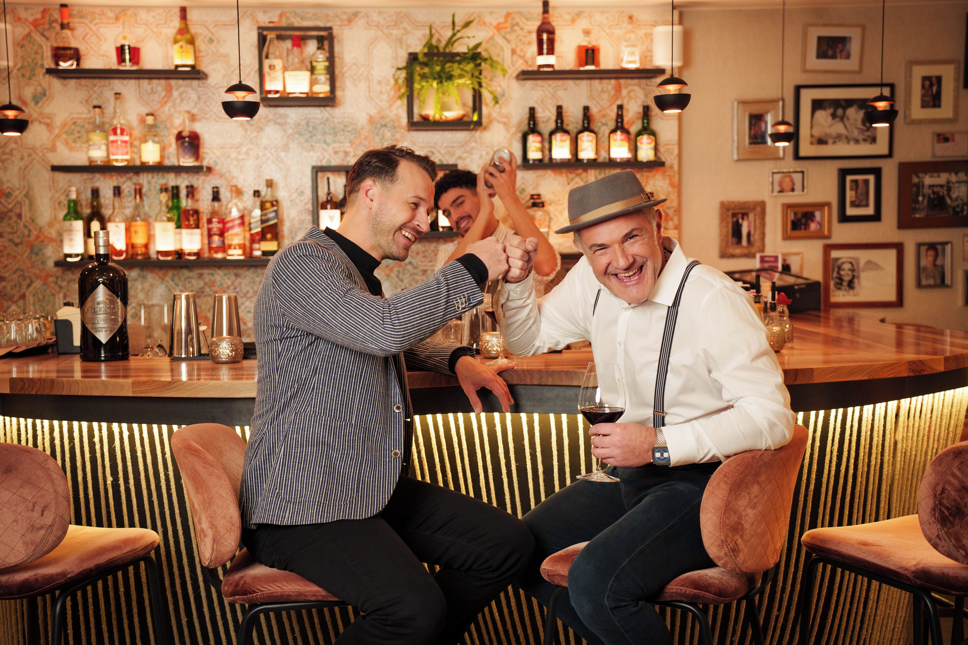 Zona de bar con varias botellas y copas en estanterías, una barra y asientos acolchados.