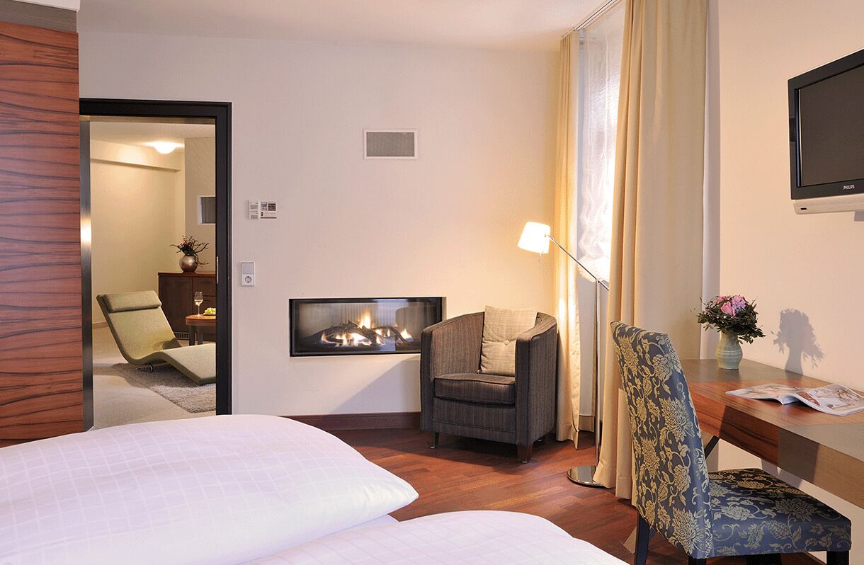 A hotel room with a bed, a chair, a desk with a lamp, a flat-screen TV, and a fireplace.