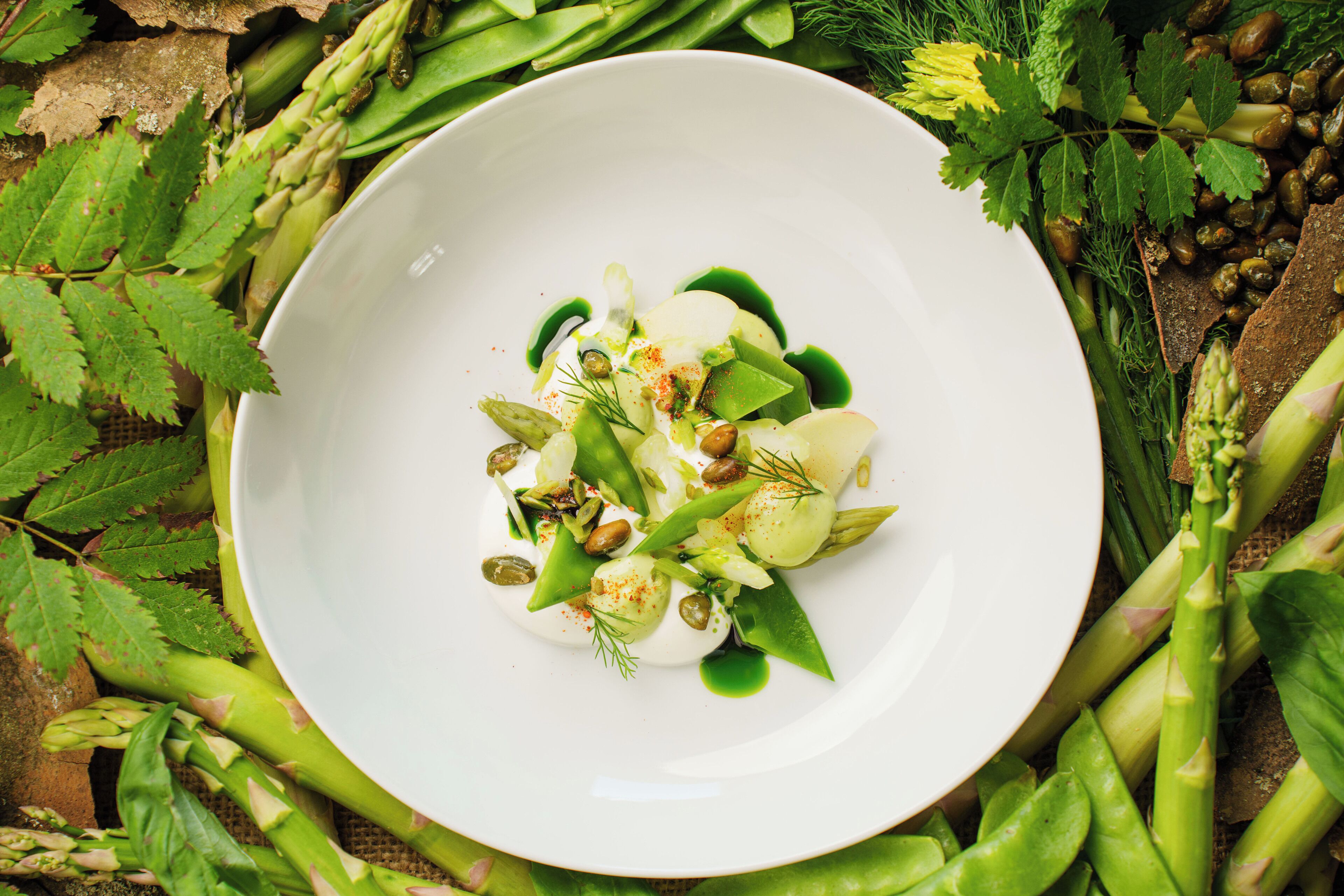 A white plate with a green vegetable dish, surrounded by asparagus and other greens.