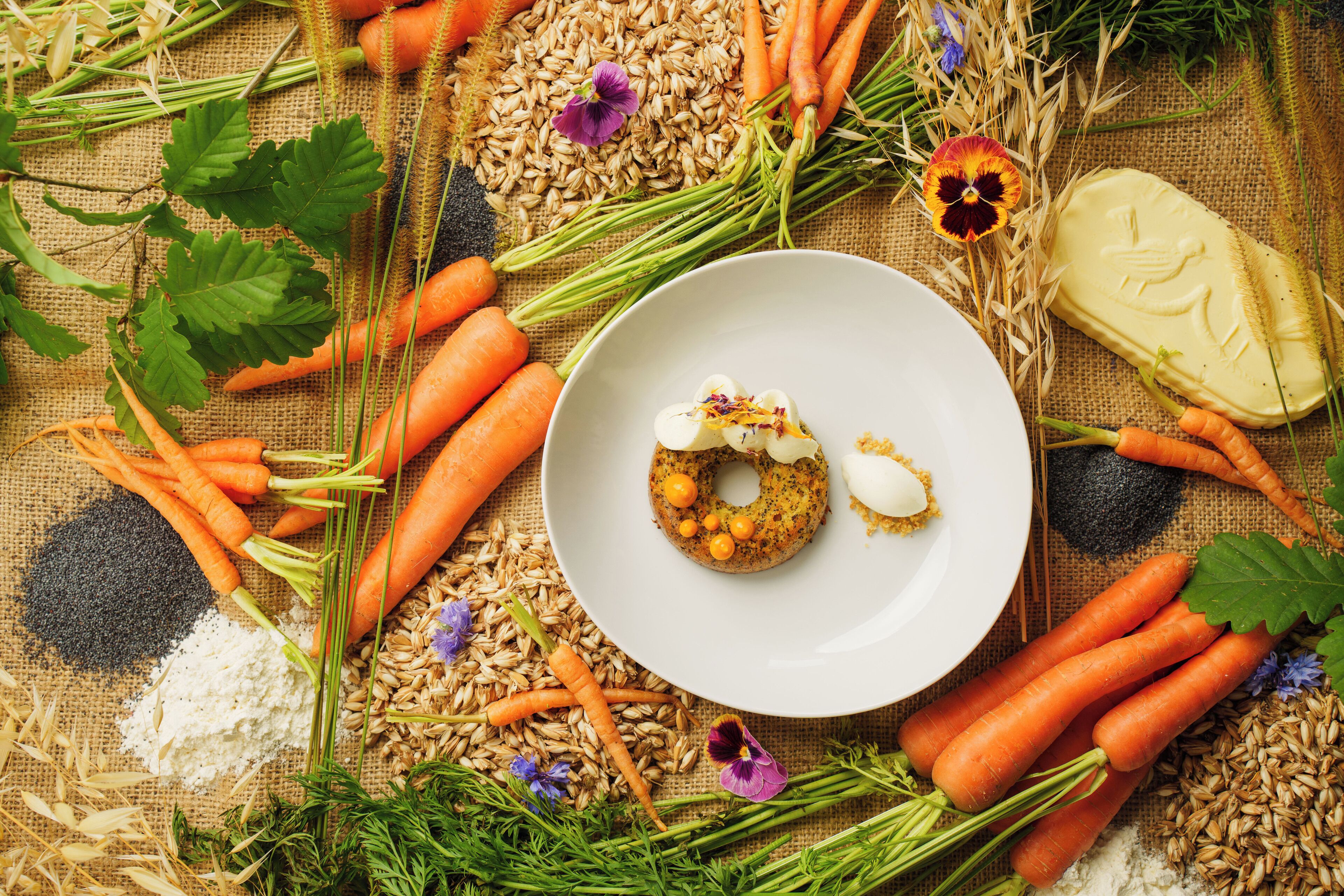 A plate with a food item, surrounded by carrots, grains, and flowers on a burlap background.