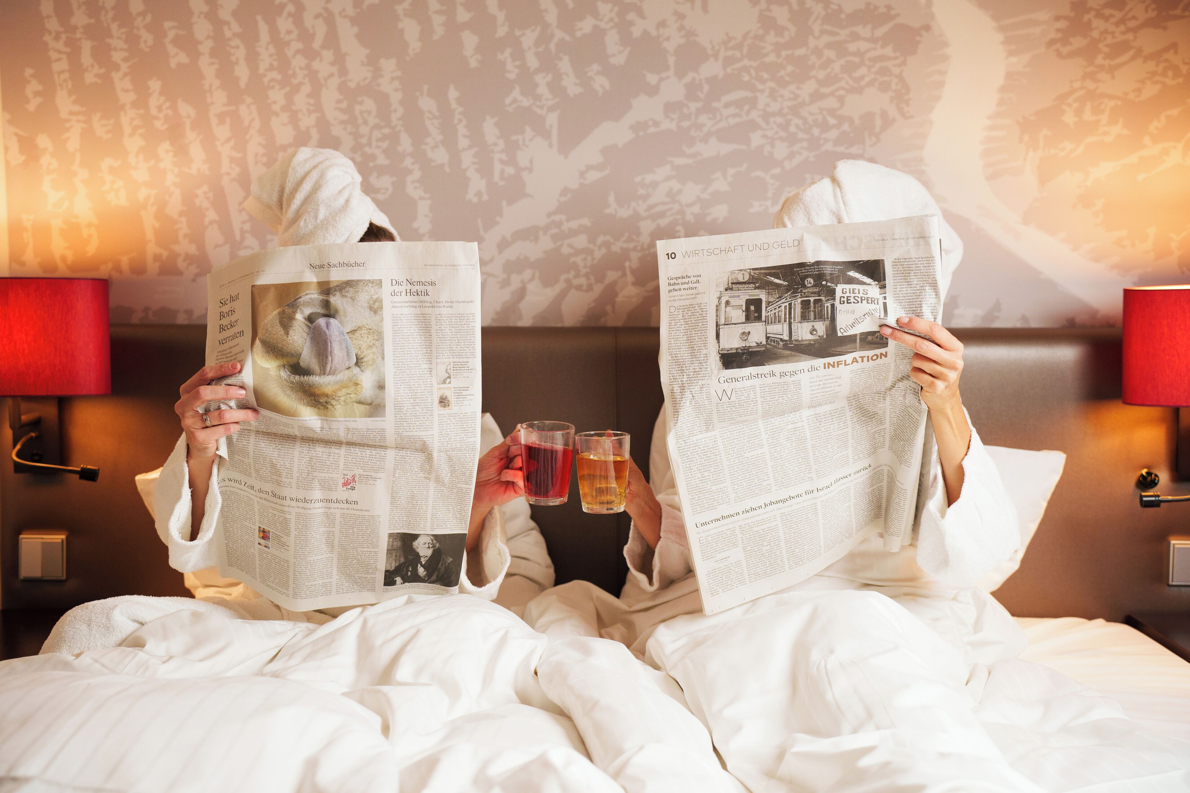 Two people reading newspapers in bed with a patterned wallpaper background and red lamps.