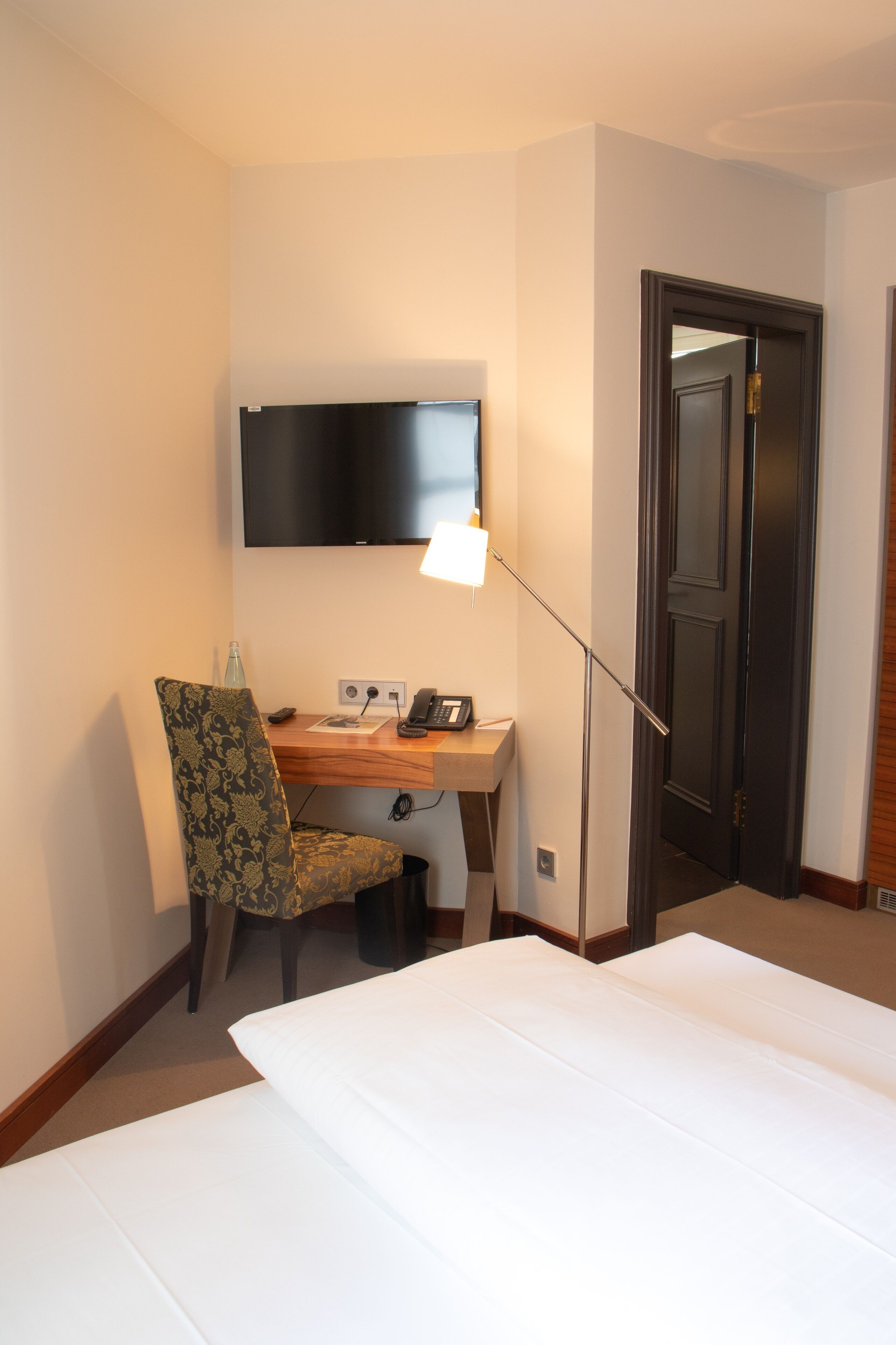 A hotel room with a bed, a desk with a chair, a flat-screen TV mounted on the wall, and a door.