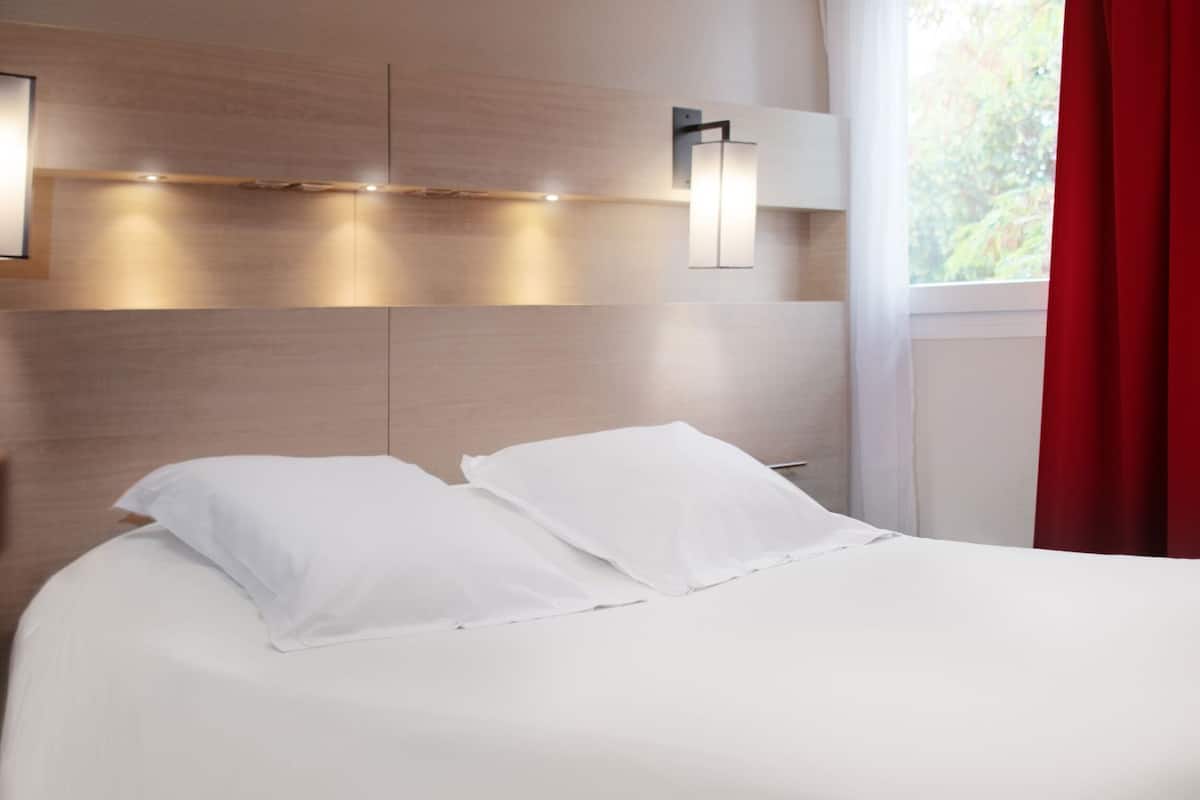 Una cama bien tendida con sábanas blancas en una habitación con cabecera de madera, una lámpara de pared y una ventana con cortinas rojas.