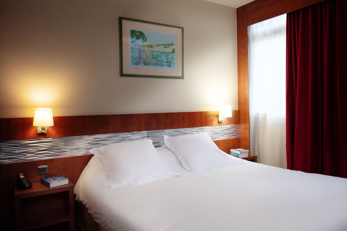 Una habitación de hotel con una cama bien hecha, dos lámparas de noche, una mesita de noche con un teléfono y un libro, y un cuadro en la pared.