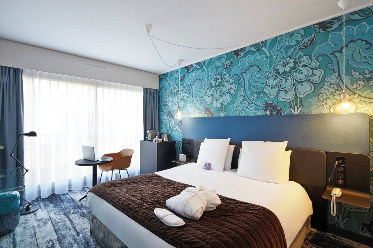 A hotel room with a bed, a desk, a chair, a desk lamp, a telephone, and a wall with a floral pattern.