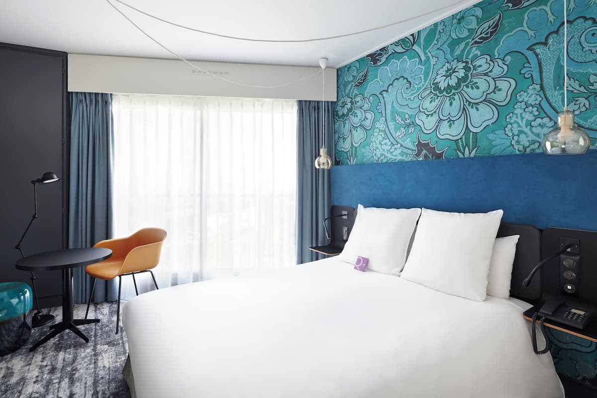 A hotel room with a large bed, a desk, a chair, a desk lamp, a telephone, and a window with curtains.