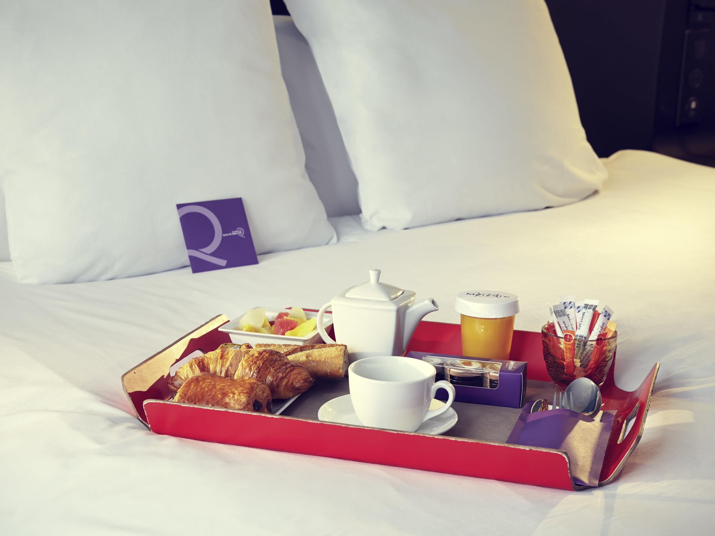 A neatly arranged breakfast tray on a bed with white linens, including croissants, a teapot, a cup, and a glass with a straw.