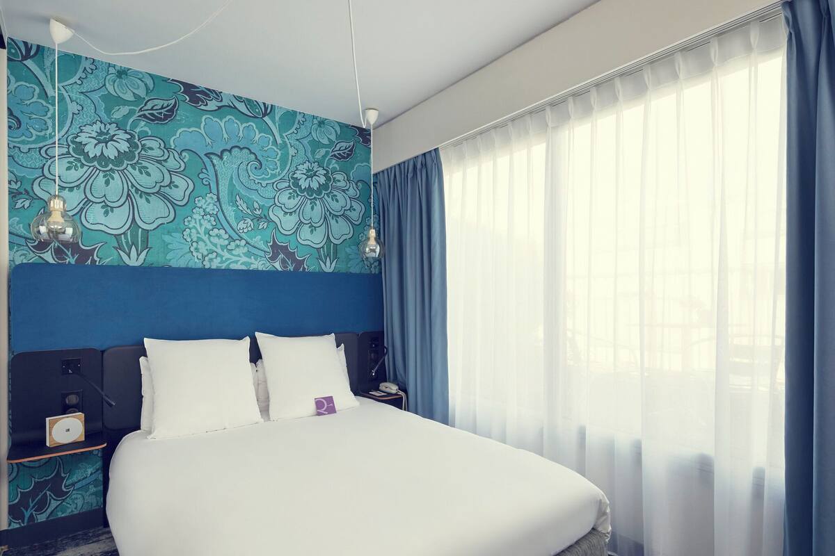 A hotel room with a bed, a nightstand, a lamp, and a window with curtains.