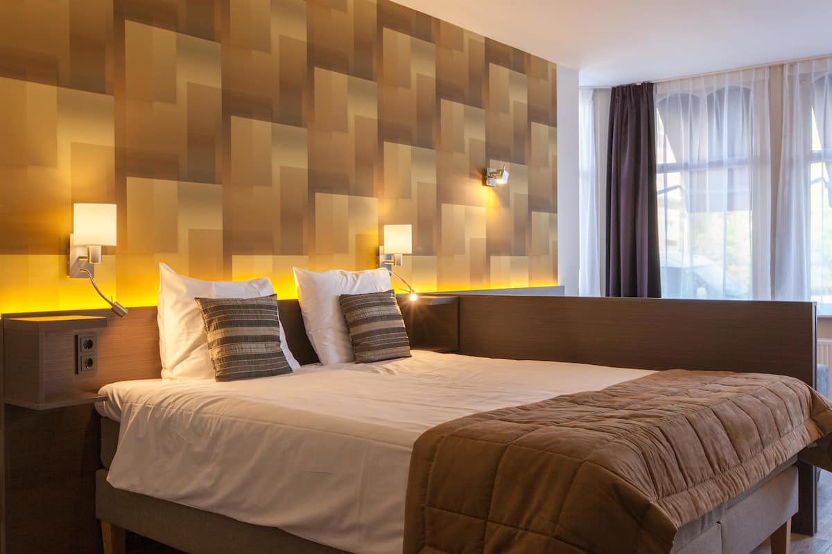 Una habitación de hotel con una cama grande, dos mesitas de noche, lámparas fijadas a la pared y un ventanal con cortinas.