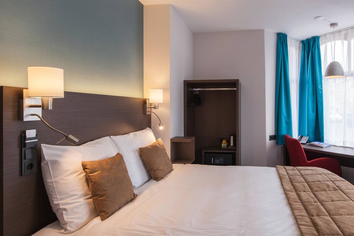 Habitación de hotel con una cama grande, lámparas de noche, un escritorio con silla, un armario y una ventana con cortinas.