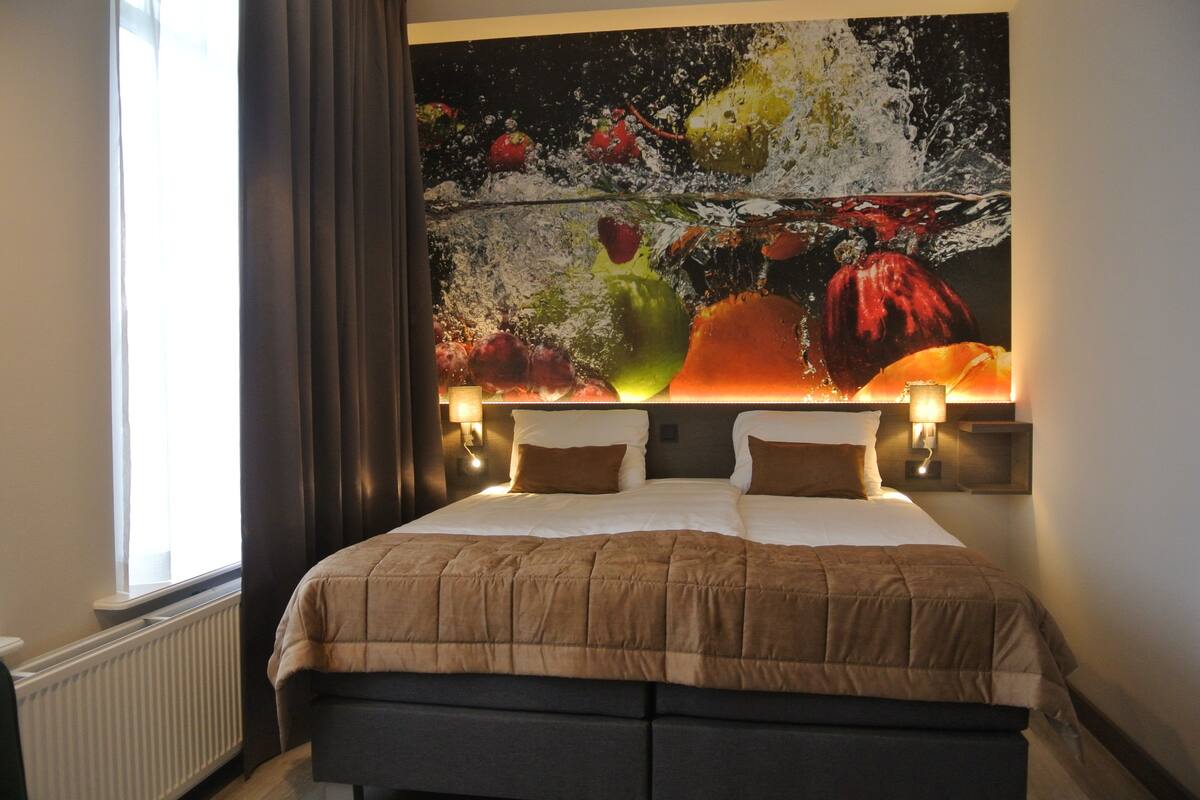 Habitación de hotel con una cama grande, cabecera con diseño de salpicadura de frutas y agua, dos lámparas de noche y una ventana con cortinas.