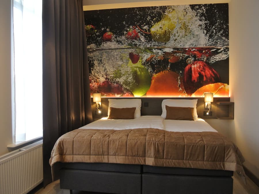 Une chambre d’hôtel avec un grand lit, une tête de lit ornée d’une œuvre d’art représentant des fruits et des gouttes d’eau, deux lampes de chevet et une fenêtre avec des rideaux.
