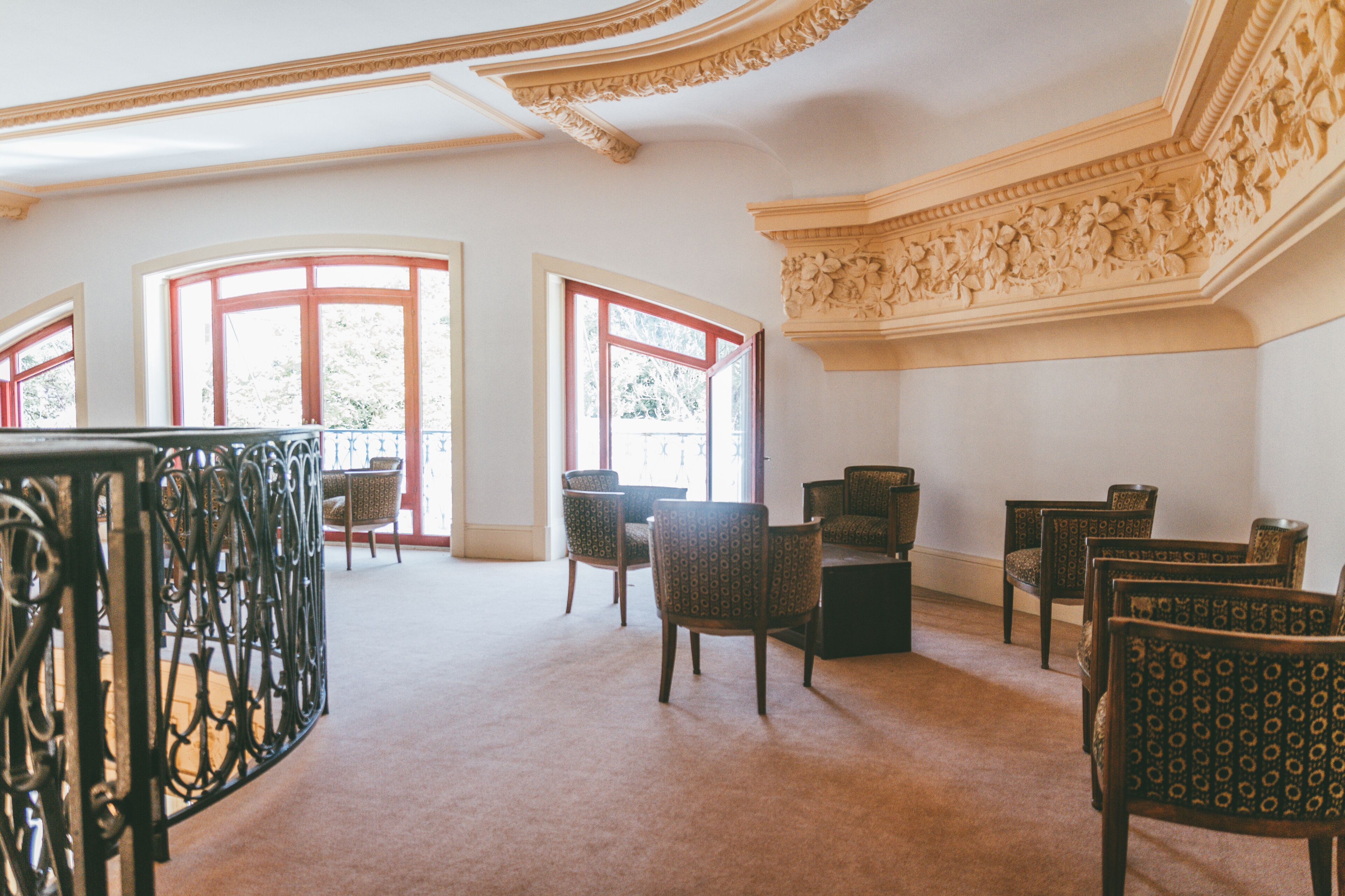 Une pièce spacieuse avec un plafond haut, des moulures décoratives et de grandes fenêtres à cadre rouge. Plusieurs fauteuils rembourrés sont disposés autour d’une table centrale.
