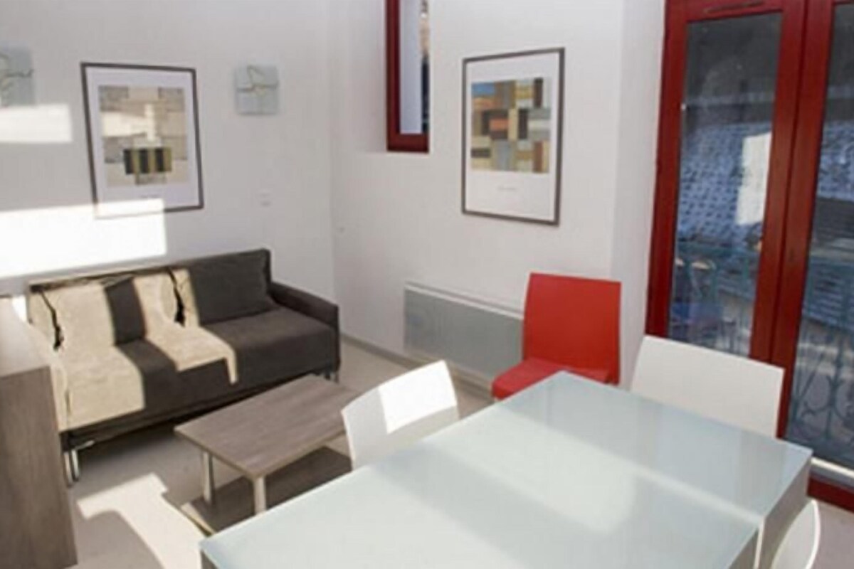A modern living room with a sofa, a dining table, and a red chair. There are framed pictures on the walls and a window with a view.